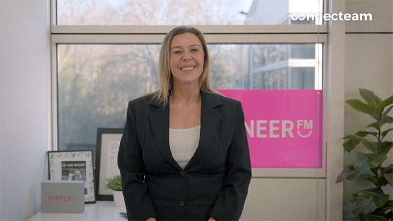 A professional shot of a woman with blonde hair, wearing a black blazer and white top, smiling in an office setting with a large window and a pink Pioneer FM sign in the background.